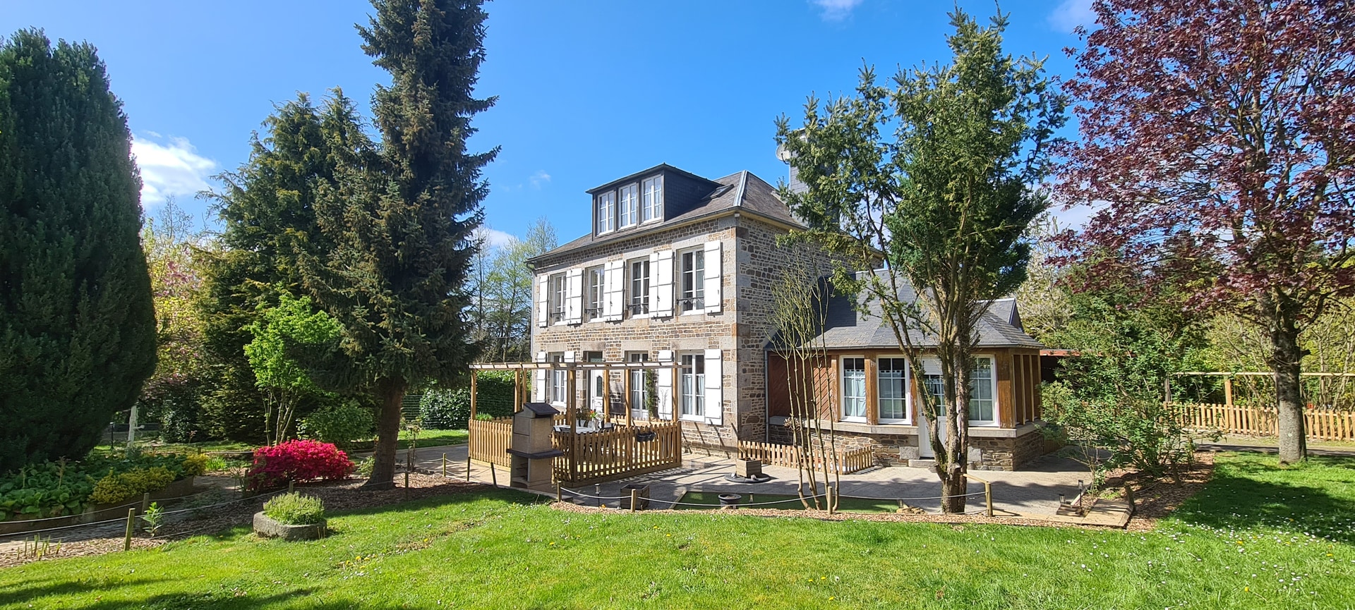 Façade en pierre de La Normandine entourée de végétation et de pelouse.
